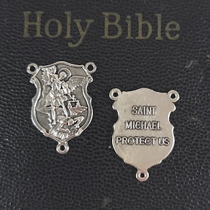 May include: Two silver-colored Saint Michael shield charms. One charm depicts Saint Michael slaying a demon. The other charm has the text "SAINT MICHAEL PROTECT US". The charms are on a dark background with the words "Holy Bible" visible.