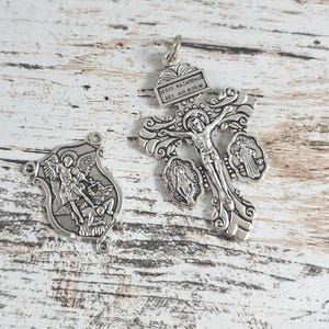 May include: Two silver religious pendants. One is a detailed crucifix with the inscription "JESUS NAZARENUS REX JUD.AEORUM". The other is a shield-shaped pendant depicting a saint. Both pendants have ornate designs and are on a light wood surface.