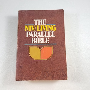 May include: The NIV/Living Parallel Bible book cover is brown with a textured surface. The title is printed in yellow and blue. The book cover features a stylized image of two leaves in yellow and orange.