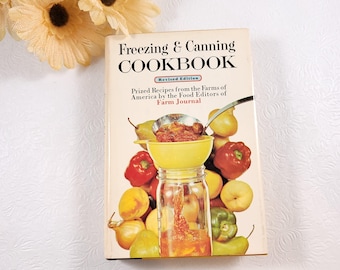 May include: A vintage cookbook titled "Freezing & Canning Cookbook" with a revised edition label. The cover features a yellow funnel with red sauce being poured into a glass jar. The background is a colorful assortment of fruits and vegetables, including pears, peaches, peppers, and onions.