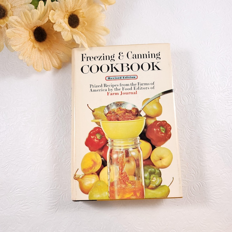 May include: A vintage cookbook titled "Freezing & Canning Cookbook" with a revised edition label. The cover features a yellow funnel with red sauce being poured into a glass jar. The background is a colorful assortment of fruits and vegetables, including pears, peaches, peppers, and onions.