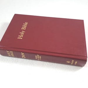 May include: A closed, burgundy-colored Holy Bible with gold lettering. The title "Holy Bible" is prominently displayed on the cover. The spine includes additional text and a cross symbol. The book is rectangular and appears to be in good condition.