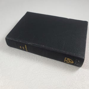 May include: A black hardcover book titled "The New Testament" with gold lettering and a gold emblem. The book has a textured cover and is lying on a white surface.