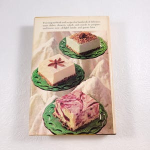 May include: A vintage cookbook cover with a green background and three different desserts on plates. The desserts are a white cheesecake with a brown crust, a green cheesecake with a white topping, and a pink and white cheesecake with a brown crust. The text on the cover reads "Freezing methods and recipes for hundreds of delicious main dishes, desserts, salads, and snacks to prepare and-freeze now-delight family and guests later."