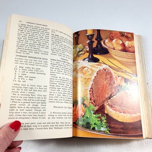 May include: Open cookbook with a vintage illustration of a roast beef dish, surrounded by vegetables and a side of rolls. The page shows a recipe for Overnight Refrigerator Rolls.