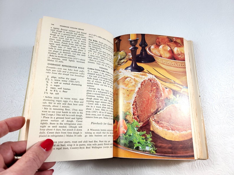 May include: Open cookbook with a vintage illustration of a roast beef dish, surrounded by vegetables and a side of rolls. The page shows a recipe for Overnight Refrigerator Rolls.