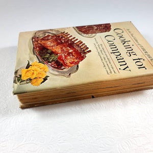 May include: A vintage cookbook titled "Cooking for Company" by the Farm Journal. The cover features a photo of a cooked rack of ribs on a silver platter with yellow roses. The book has aged, yellowed pages.