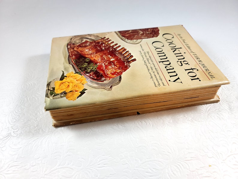 May include: A vintage cookbook titled "Cooking for Company" by the Farm Journal. The cover features a photo of a cooked rack of ribs on a silver platter with yellow roses. The book has aged, yellowed pages.
