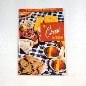 May include: A vintage cookbook titled "The Cheese Cookbook" with a red cover and a checkered blue and white tablecloth background. The cover features a variety of cheeses, sandwiches, and a dish of tomatoes with cheese sauce.