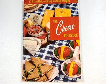 May include: A vintage cookbook titled "The Cheese Cookbook" with a red cover and a checkered blue and white tablecloth background. The cover features a variety of cheeses, sandwiches, and a dish of tomatoes with cheese sauce.