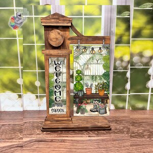 May include: Wooden welcome sign featuring a greenhouse scene and the text "Welcome to our garden". The sign has a brown wood finish and is decorated with a white heart.