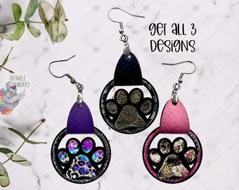 May include: Three dangle earrings featuring a circular frame with a central animal paw print cutout. The earrings have textured leather-like tabs in purple, black, and pink, with colorful leopard print patterns. Text reads: get all 3 designs.