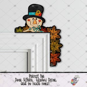 May include: A fall-themed door or window decor featuring a scarecrow with a black top hat, a blue bow, and a face with a stitched smile. The scarecrow is surrounded by colorful autumn leaves.