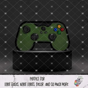Könnte beinhalten: Ein grünes Videospiel-Controller-Design auf einem schwarzen Sockel. Der Controller hat farbige Tasten und ein Steuerkreuz. Der Text unten lautet: "PERFECT FOR LIGHT BASES, NIGHT LIGHTS, DECOR, AND SO MUCH MORE."