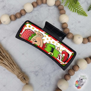 May include: A black car vent clip with a Christmas elf design. The elf is wearing a green suit with a red hat and white trim. The background is red and white with gold polka dots.