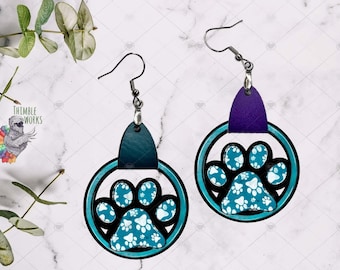 May include: A pair of dangle earrings featuring circular frames with a teal and black paw print design. The left earring has a dark teal leather-like tab, and the right has a purple tab. Both hang from silver-toned metal hooks.