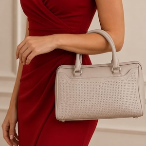 May include: A light gray leather handbag featuring a rectangular silhouette with rounded corners, dual top handles, and a perforated front panel. The bag includes silver-toned hardware attachments for the handles and a smooth leather trim along the top edge.