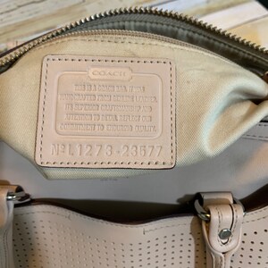 May include: A beige leather handbag featuring a perforated exterior and a cream-colored interior lining. A rectangular leather patch is sewn inside, embossed with the Coach logo and the text: This is a Coach bag. It was handcrafted from genuine leather. Its superior craftsmanship and attention to detail reflect our commitment to enduring quality. No L 1273-23577.