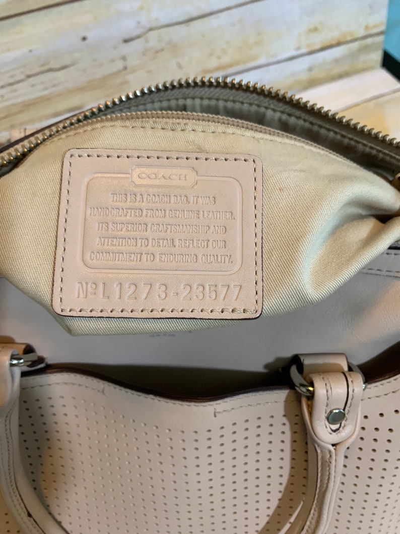 May include: A beige leather handbag featuring a perforated exterior and a cream-colored interior lining. A rectangular leather patch is sewn inside, embossed with the Coach logo and the text: This is a Coach bag. It was handcrafted from genuine leather. Its superior craftsmanship and attention to detail reflect our commitment to enduring quality. No L 1273-23577.