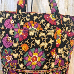 Op de afbeelding: Zwarte tote bag met een levendig bloemenpatroon in tinten roze, paars, geel en oranje. De tas heeft gewatteerde stiksels en stevige handvatten. Het bloemmotief staat tegen een zwarte achtergrond.