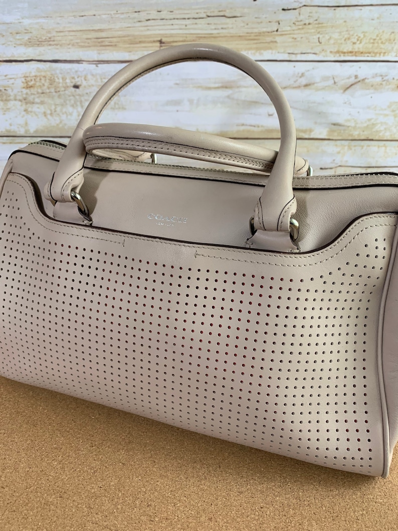 May include: A light beige leather handbag featuring a perforated front panel with small circular cutouts. The bag has two rounded top handles, silver-toned hardware, and the word COACH NEW YORK embossed in small silver lettering on the front.