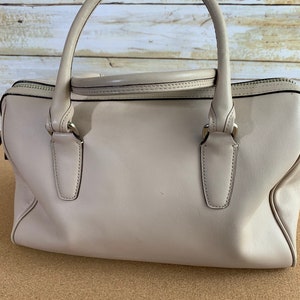 May include: A light beige leather handbag featuring a structured rectangular body, two rounded top handles, and a top zipper closure. The bag includes silver-toned metal hardware attachments for the handles and a smooth, solid-colored exterior finish.