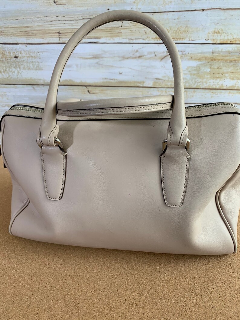 May include: A light beige leather handbag featuring a structured rectangular body, two rounded top handles, and a top zipper closure. The bag includes silver-toned metal hardware attachments for the handles and a smooth, solid-colored exterior finish.