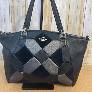 以下が含まれることがあります： 前面に幾何学的なパッチワークデザインが施された黒いレザートートバッグ。バッグは、シルバーのジッパー、デュアルハンドル、コーチのロゴが特徴です。小さなタグがハンドルからぶら下がっています。バッグは革とスエード製です。