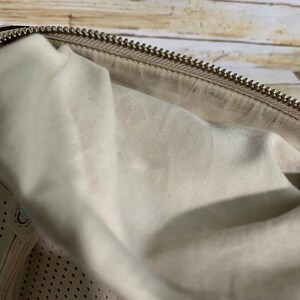 May include: A beige handbag interior featuring a light-colored fabric lining and a metallic zipper closure along the top edge. The lower section of the bag displays a perforated pattern on the beige material, showing a consistent texture throughout.