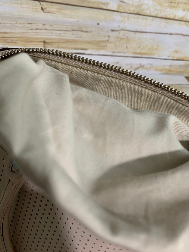 May include: A beige handbag interior featuring a light-colored fabric lining and a metallic zipper closure along the top edge. The lower section of the bag displays a perforated pattern on the beige material, showing a consistent texture throughout.