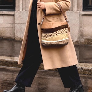 May include: A shoulder bag featuring a multi-textured design with wavy horizontal panels in shades of tan, dark brown, and gold. The bag includes sections with a repeating circular logo pattern, a metallic gold strip, and a dark brown leather-like base.