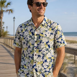 Peut inclure: Une chemise à manches courtes boutonnée avec un imprimé tropical vibrant composé de fleurs d'hibiscus blanches, de feuilles de monstera vertes et d'ananas jaunes sur un fond bleu marine profond, dotée d'un col classique et de boutons marron.