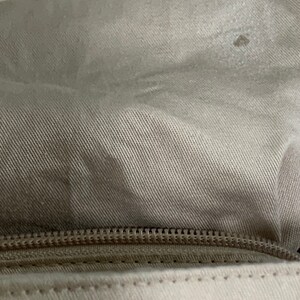 May include: A close-up view of a beige fabric pouch featuring a horizontal zipper closure. The upper section is made of a textured, light tan woven material, while the lower section consists of a smooth, off-white fabric with visible stitching.