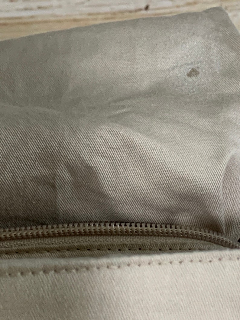 May include: A close-up view of a beige fabric pouch featuring a horizontal zipper closure. The upper section is made of a textured, light tan woven material, while the lower section consists of a smooth, off-white fabric with visible stitching.