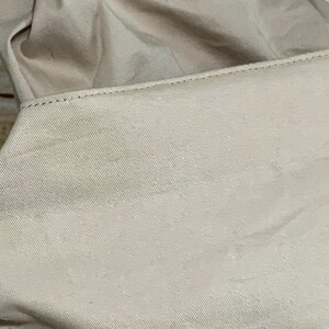 May include: A beige fabric item featuring a prominent rectangular panel with a distinct diagonal twill weave texture. The panel is attached with visible straight stitching along the top edge, while the rest of the material appears soft and folded.