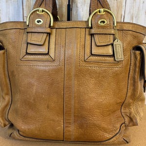 May include: A tan leather tote bag with gold-tone hardware. The bag features two top handles, multiple pockets, and a small tag with the brand name. The leather has a textured appearance with visible stitching details. The bag has a classic, structured design.