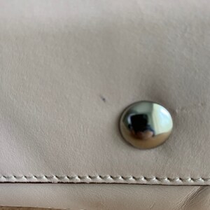 May include: A smooth, light beige leather surface featuring a single, polished, circular silver-tone metal stud. The leather is joined by a horizontal line of precise, tonal stitching that runs across the lower portion of the frame.