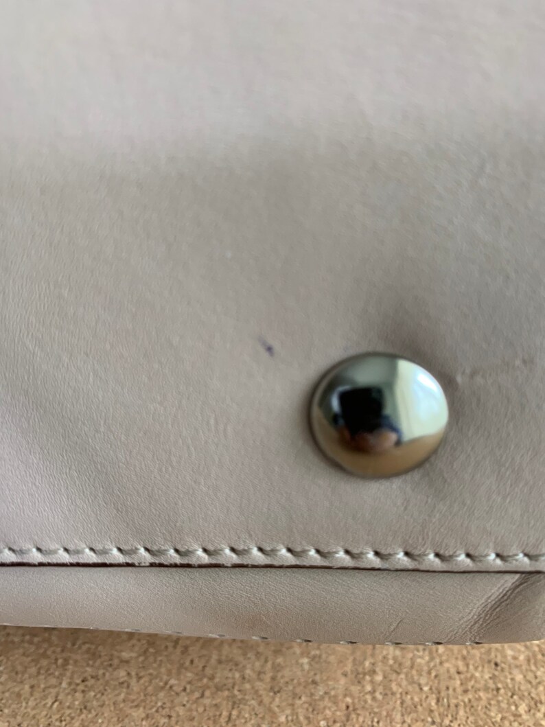 May include: A smooth, light beige leather surface featuring a single, polished, circular silver-tone metal stud. The leather is joined by a horizontal line of precise, tonal stitching that runs across the lower portion of the frame.