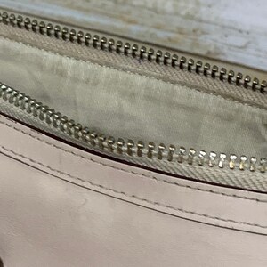May include: A light beige leather pouch featuring a silver-toned metal zipper closure. The interior is lined with a matching light beige fabric, and the exterior displays clean, precise stitching along the edge of the smooth leather material.