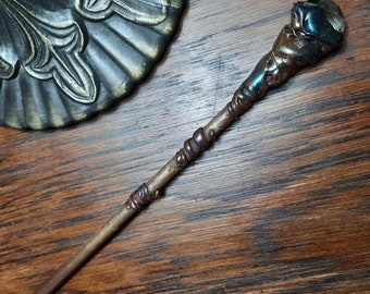 May include: A wooden wand with a iridescent blue and green crystal at the top. The wand is decorated with copper wire and purple beads.