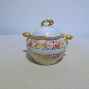 May include: A small, antique sugar bowl with a lid. The bowl is cream-colored with a floral design of pink and yellow flowers. It has gold-colored handles and a gold handle on the lid. The bowl is likely made of porcelain.