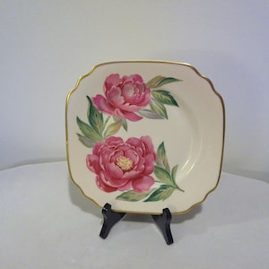 May include: A square, off-white plate with a gold trim and a floral design featuring two pink peonies and green leaves. The plate is displayed on a black stand.