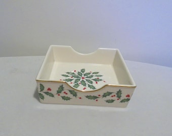 May include: A square cream-coloured ceramic napkin holder featuring a painted pattern of green holly leaves and red berries on the exterior and interior base. The top edges are finished with a thin gold trim and a central cutout.