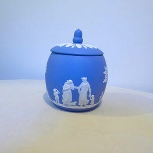 May include: A blue ceramic jar with a matching lid featuring a white acorn-shaped finial. The exterior is decorated with white relief figures of people and a small animal, while the lid edge is adorned with a white leaf pattern design.