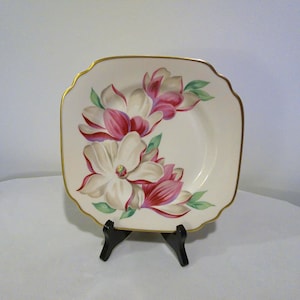 May include: A square, off-white plate with a gold rim and a floral design. The flowers are white and pink with green leaves. The plate is displayed on a black stand.
