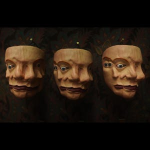 May include: Three beige, sculpted face masks arranged in a row. Each mask features multiple overlapping facial features, including several eyes and noses, with dark red lips. The masks have a textured, skin-like appearance and are mounted on a dark surface.