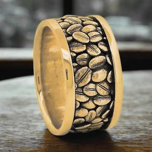 Op de afbeelding: Een goudkleurige ring met een band van gedetailleerde koffieboon-carvingen. De ring heeft een gepolijste, gladde buitenkant en een getextureerde binnenband gevuld met koffieboon-ontwerpen. De ring wordt gepresenteerd op een houten oppervlak.