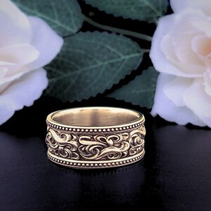 May include: A gold band ring with a detailed, intricate design of swirling patterns. The ring is set against a background of white roses and green leaves.