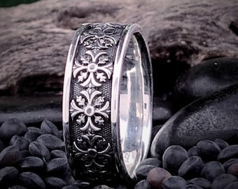 May include: A silver band ring with a detailed floral design. The ring is set against a background of black pebbles.
