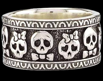 Könnte beinhalten: Ein silberner Ring mit einem Band, das mit fünf Totenkopfmotiven verziert ist. Die Totenköpfe wechseln sich ab zwischen männlich und weiblich, wobei die weiblichen Totenköpfe Schleifen tragen.